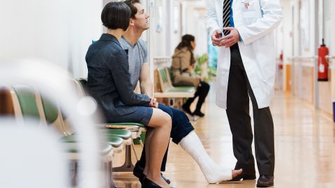 Gebrochenes Sprunggelenk (Quelle: Getty Images/TommL) Ein Mann mit Gips am Bein und eine Frau sitzen in einem Wartebereich. Neben ihnen steht ein Arzt. Sie sprechen miteinander.
