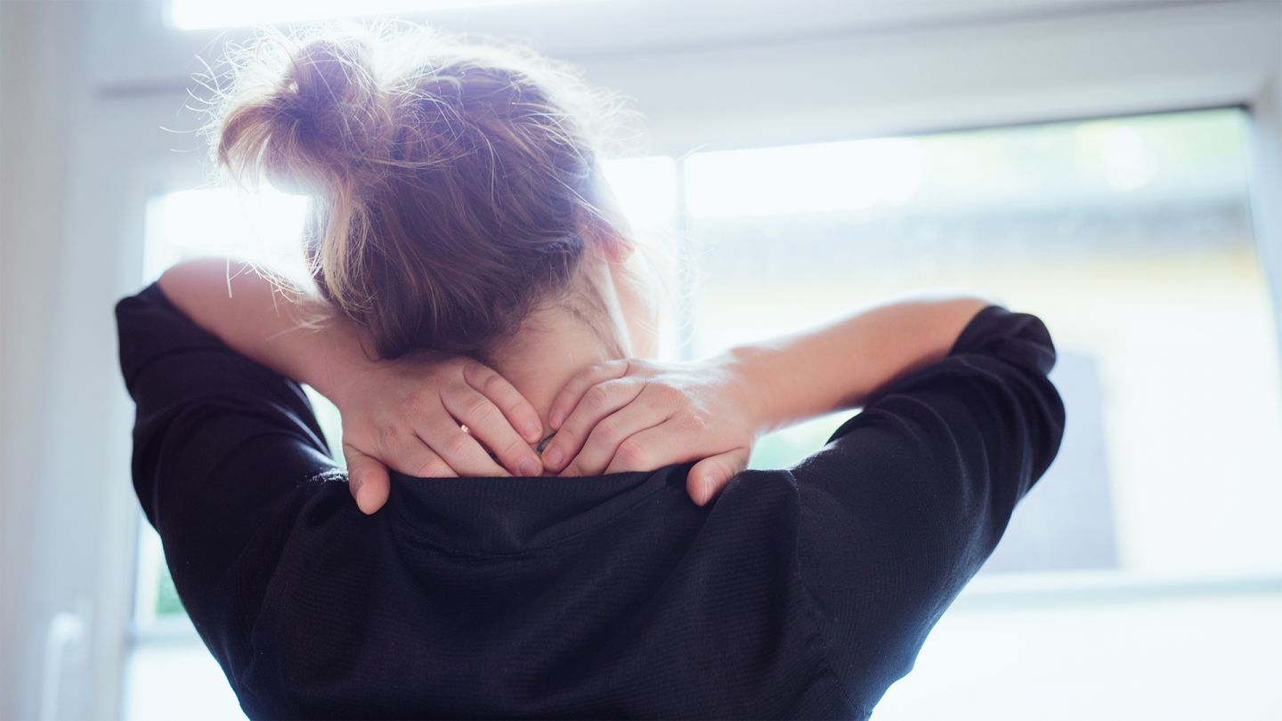 Stress (Quelle: Getty Images/Guido Mieth) Stress: Eine Frau mit hochgebundenen Haaren steht vor dem Fenster und streckt die Arme nach oben und drückt mit den Händen gegen ihren verspannten Nacken. Sie hält den Kopf dabei schräg in Richtung Schulter, um ihren Nacken zu dehnen.
