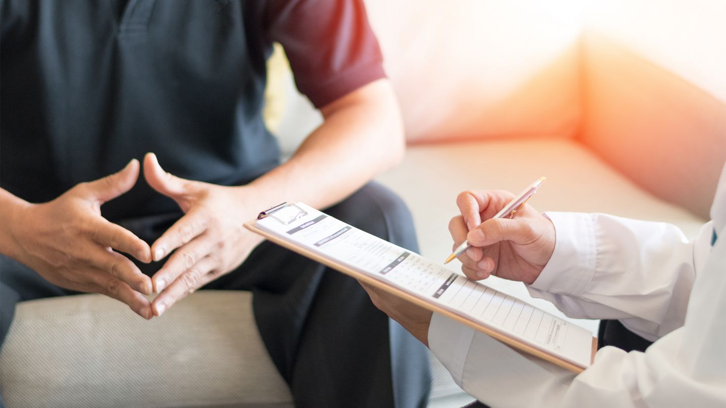 Syphilis (Quelle: Getty Images/Pornpak Khunatorn) Syphilis: Ein Mann sitzt auf einer Couch, seine Hände Formen eine Raute. Schräg vor ihm sitzt ein Arzt, der ein Klemmbrett und einen Kugelschreiber in den Händen hält.