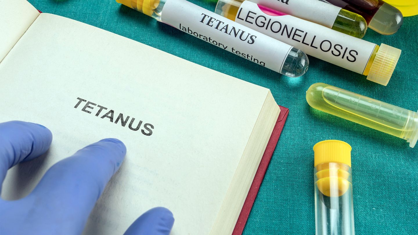 Tetanus (Quelle: Getty Images/digicomphoto) Eine Behandschuhte Hand zeigt auf den Begriff Tetanus in einem aufgeschlagenem Buch. Daneben liegen einige Reagenzgläsern mit farbigen Flüssigkeiten.