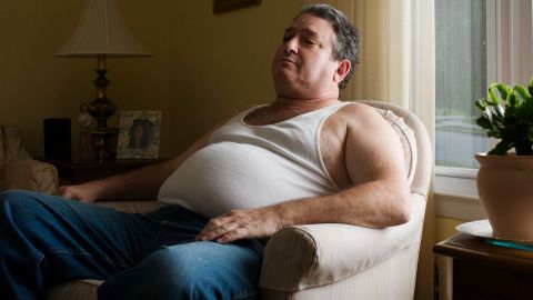 Übergewichtiger Mann (Symbolbild) (Quelle: Getty Images/Michael Greenberg) Ein übergewichtiger Mann sitzt in einem Sessel. Unter dem Feinrippunterhemd, das der Mann trägt, ist ein dicker Bauchansatz zu erkennen.