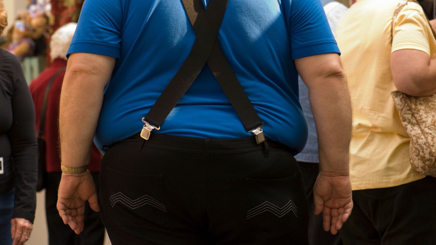 Mann mit Übergewicht (Quelle: Getty Images/JPecha) Übergewicht und Adipositas: Ein Mann, der Latzhose trägt, steht in einer Menschenmenge. Links und rechts am Hosenbund drückt der Bauch über den Hosenrand.