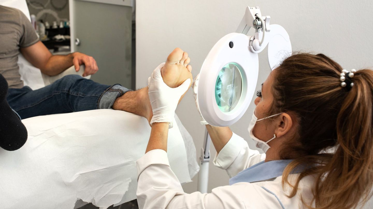 Warzen-Behandlung (Quelle: Getty Images/pankration) Warzen: Ein Mann sitzt auf einem Behandlungsstuhl in einer Praxis und streckt ein Bein nach vorne. Eine junge Frau, die einen weißen Kittel, Mundschutz und Medizinhandschuhe trägt, hält einen Fuß des Mannes fest und trägt Stickstoff auf eine Stelle auf.