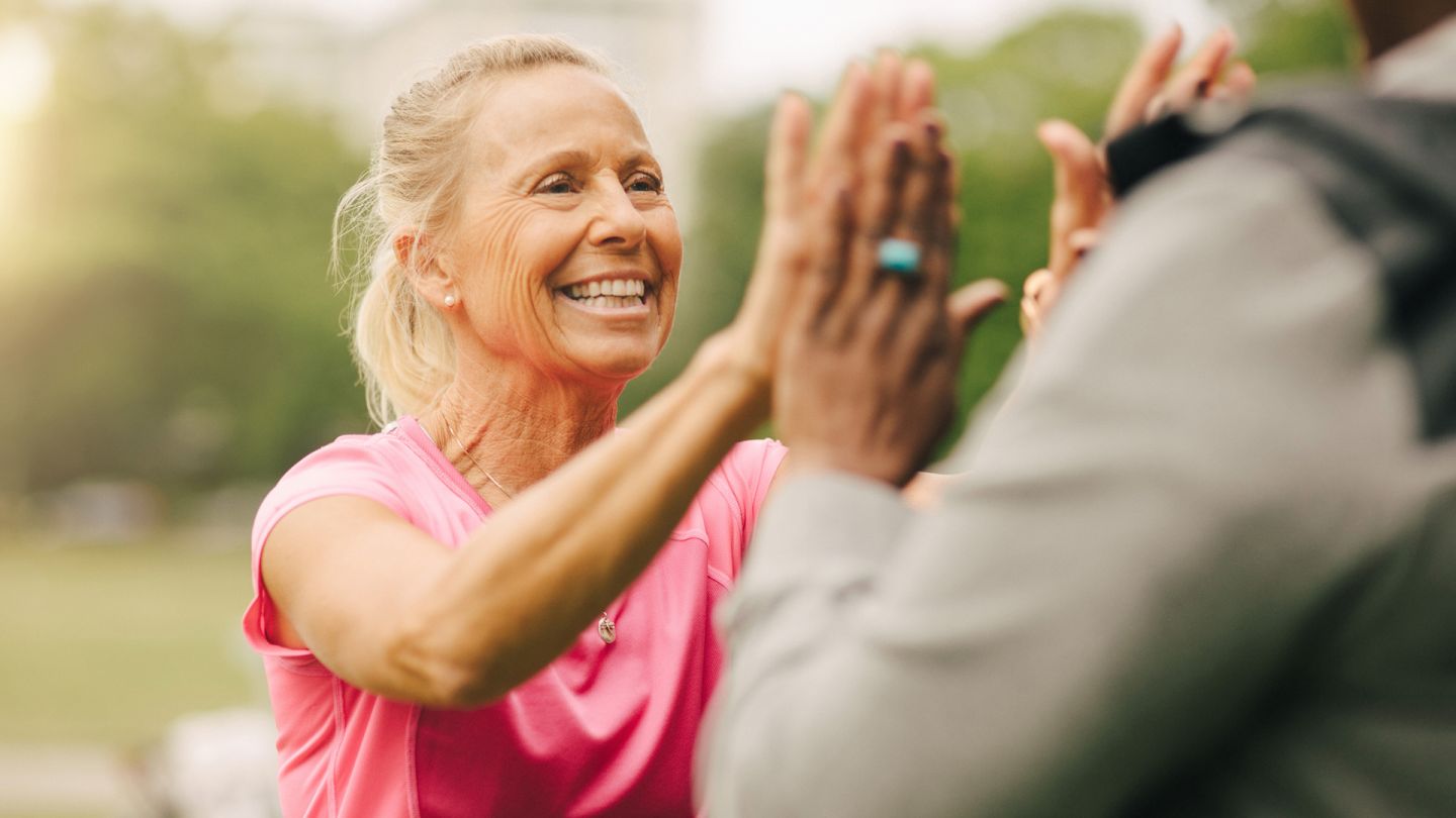 Sportliche Frau (Quelle: Getty Images/Maskot) Wechseljahre: Eine Frau mittleren Alters trägt sportliche Kleidung und klatscht ihre Handflächen in die Handflächen der anderen Frau.