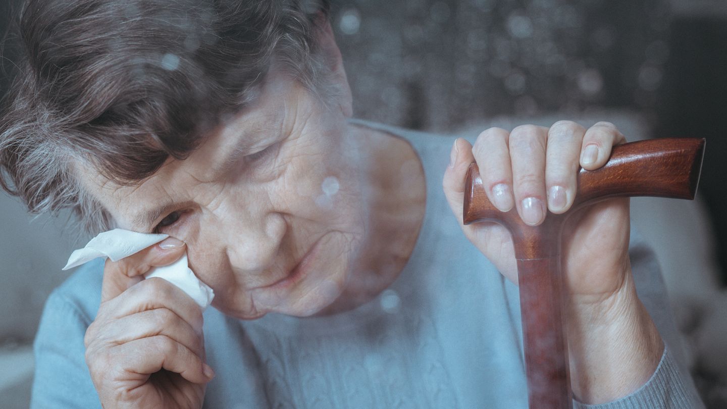 Frau mit Alzheimer-Demenz (Symbolbild) (Quelle: Getty Images/KatarzynaBialasiewicz) Alzheimer-Demenz: Eine Frau mit Gehstock hält ein Taschentuch an ihr Auge und blickt nach unten.