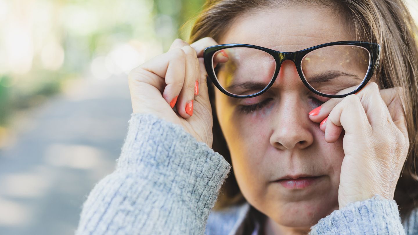 Eine Frau reibt sich das Auge (Quelle: Getty Images/Madrolly) Augenlidentzündung: Eine Frau, die eine Brille trägt, schiebt die Brille mit einer Hand leicht nach oben und fasst sich mit der anderen Hand ans geschlossene Auge.