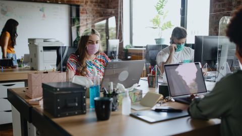 Coronavirus Vorbeugung (Quelle: iStock.com/Anchiy) Zwei Frauen und ein Mann sitzen gemeinsam an einem großen Schreibtisch und arbeiten. Sie alle tragen einen Mund-Nase-Schutz und sind durch Plexiglasscheiben voneinander getrennt.