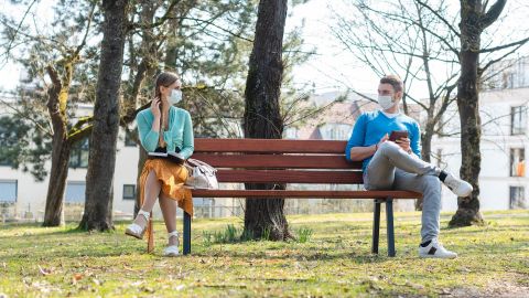 Abstand halten mit Atemschutzmasken (Quelle: Getty Images/kzenon) Ein Mann und eine Frau sitzen auf einer Parkbank und gucken sich an. Zwischen ihnen ist ungefähr 1,5 Meter Abstand. Beide tragen einen Mundschutz.