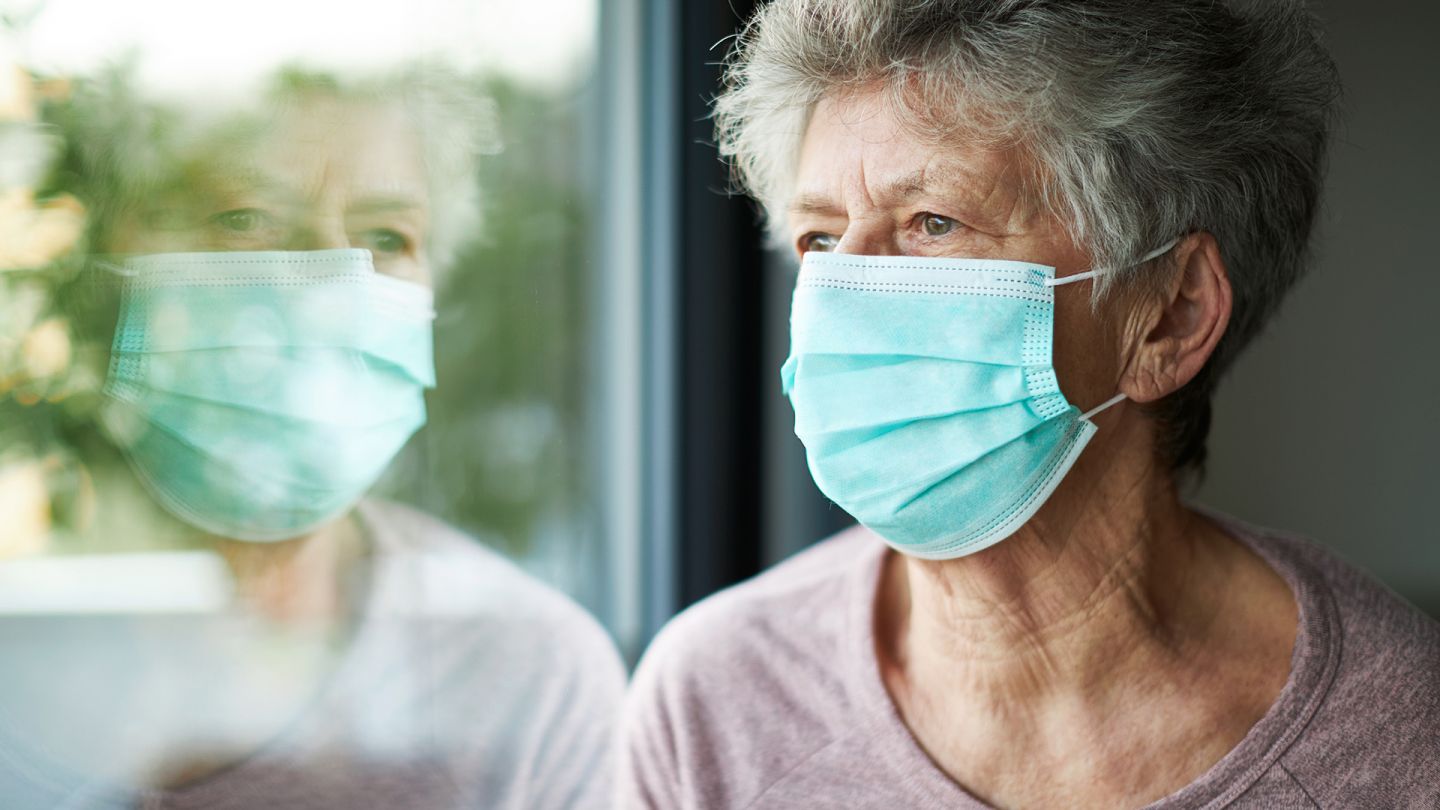 Frau mit Mundschutz (Symbolbild) (Quelle: Getty Images/Kai und Kristin Fotografie) COVID-19: Eine ältere Frau schaut aus dem Fenster. Über Mund und Nase trägt sie einen Mundschutz.