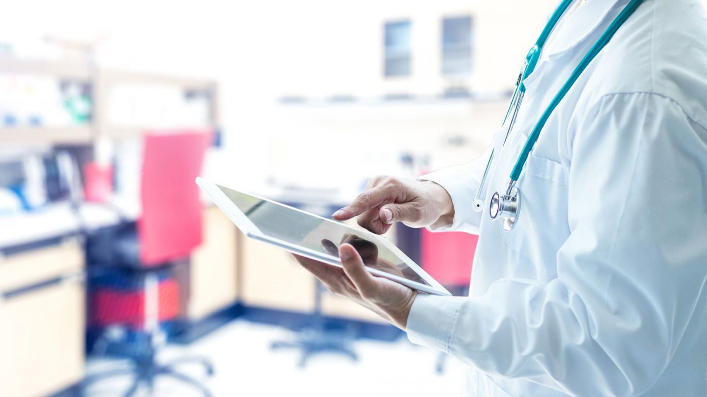 Arzt mit Tablet (Quelle: Getty Images/Pornpak Khunatorn) Elektronische Patientenakte: Ein Arzt steht in einer Praxis und hält ein Tablet in der linken Hand. Mit dem Zeigefinger der rechten Hand berührt er den Bildschirm.