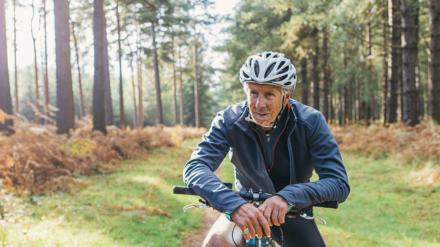 Ernährung und Bewegung im Alter (Quelle: Getty Images/Justin Paget) Ernährung und Bewegung im Alter: Ein älterer Mann in Fahrradkleidung pausiert während einer Radtour im Wald und stützt sich dabei auf den Lenker seines Fahrrads.
