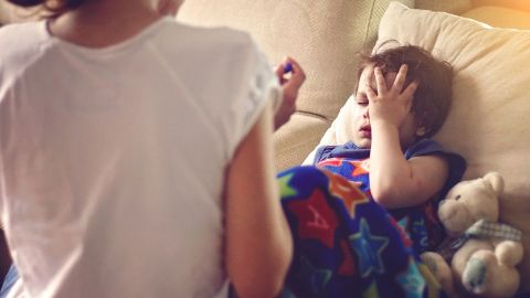 Fieber bei Kindern - Diagnostik (Quelle: Getty Images/anandaBGD) Fieber? Ein Kind, das erschöpft aussieht, liegt auf einer Couch und fasst sich an den Kopf.
