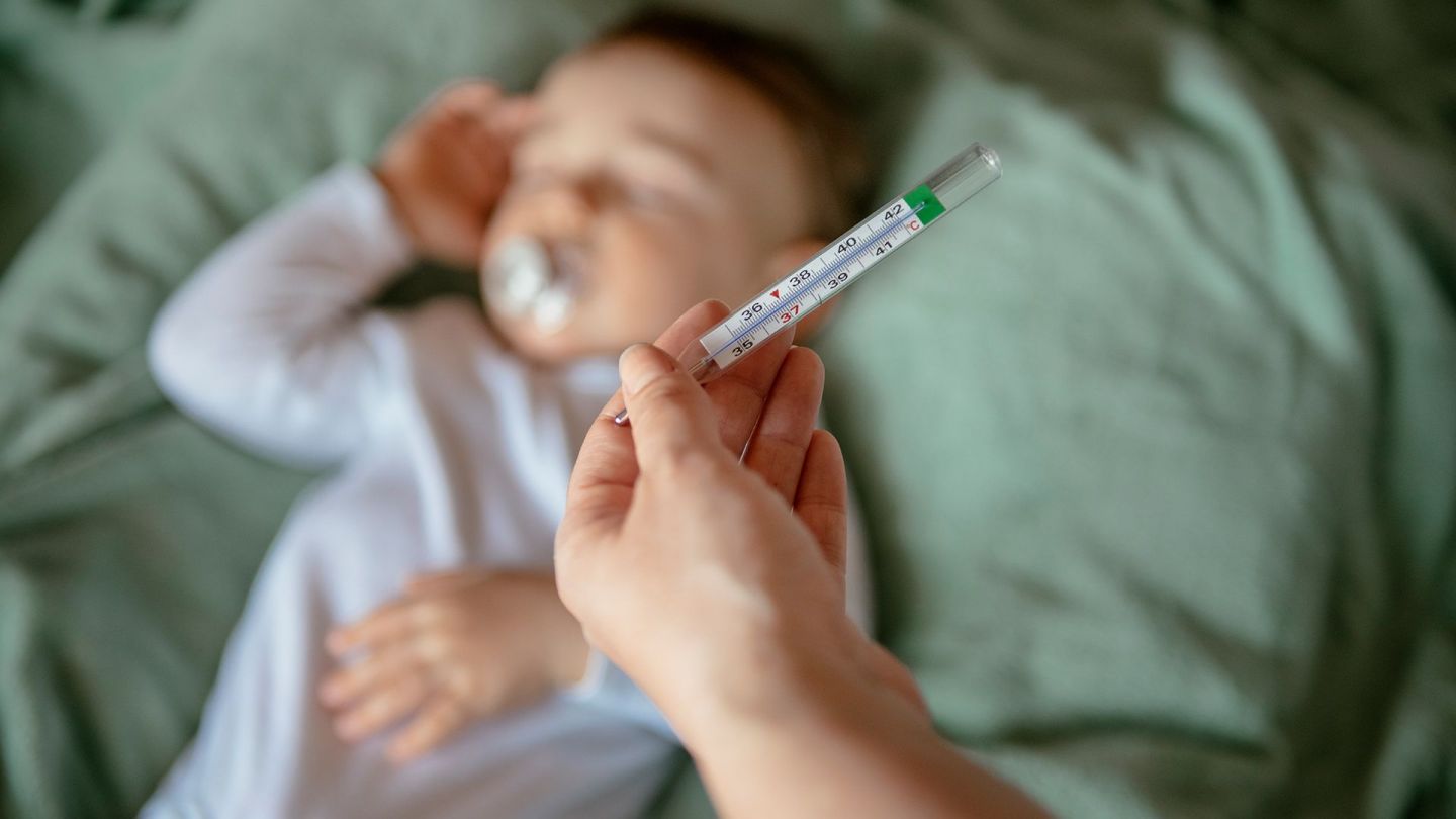 Fieber messen bei Kindern (Quelle: Getty Images/Milos Dimic) Fieber bei Kindern: Eine Hand hält ein Fieberthermometer, im Hintergrund liegt ein Baby in einem Bett.