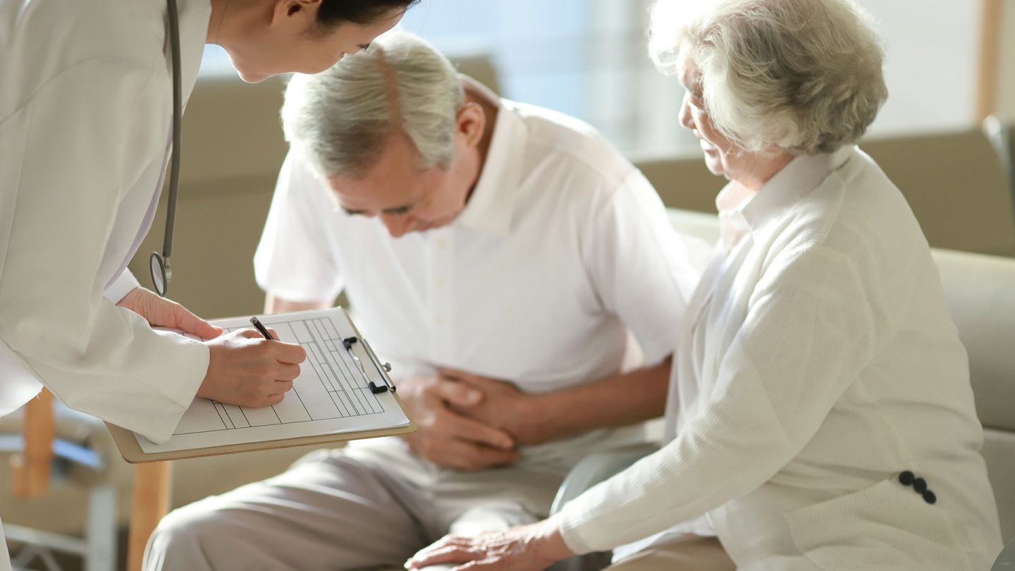 Mann mit Bauchschmerzen (Quelle: Getty Images/Runstudio) Gallensteine: Eine älterer Mann sitzt auf einer Couch, hat seinen Oberkörper nach vorne gebeugt und presst beide Hände an den Bauch. Eine Frau sitzt neben ihm und berührt mit einer Hand sein linkes Bein. Vor ihnen steht ein Arzt, der sich Notizen macht.