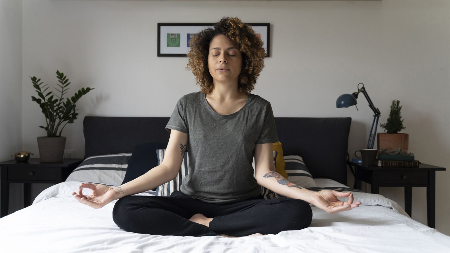 Gesund mit Entspannung (Quelle: Getty Images/ Westend61) Gesund mit Entspannung: Eine Frau sitzt auf ihrem Bett im Schneidersitz und meditiert. Sie hat beide Hände auf ihren Knien abgelegt und die Hände in Chinmudra-Haltung. Sie hat die Augen geschlossen und atmet aus.