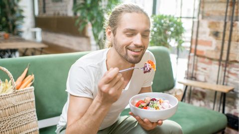Gesunde Ernährung (Quelle: Getty Images/Ross Helen) Ein junger Mann sitzt auf einem Sofa und isst einen Salat. Er lächelt dabei. Hinter ihm steht auf dem Sofa ein Korb mit Gemüse.