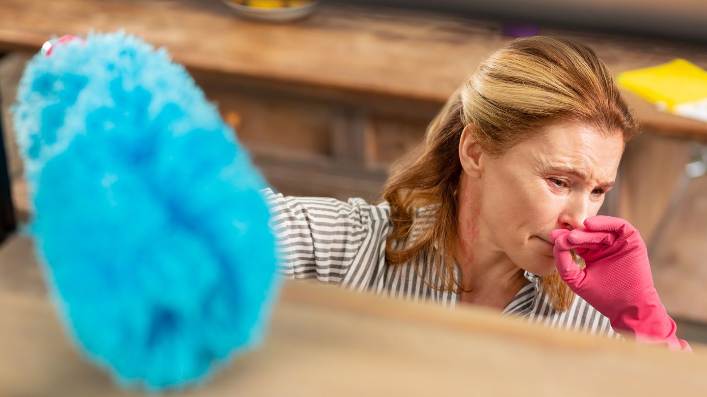 Hausstauballergie (Symbolbild) (Quelle: Getty Images/Zinkevych) Hausstauballergie: Eine Frau wischt Staub und hält sich die Hand ins Gesicht. Ihre Augen sind gerötet.