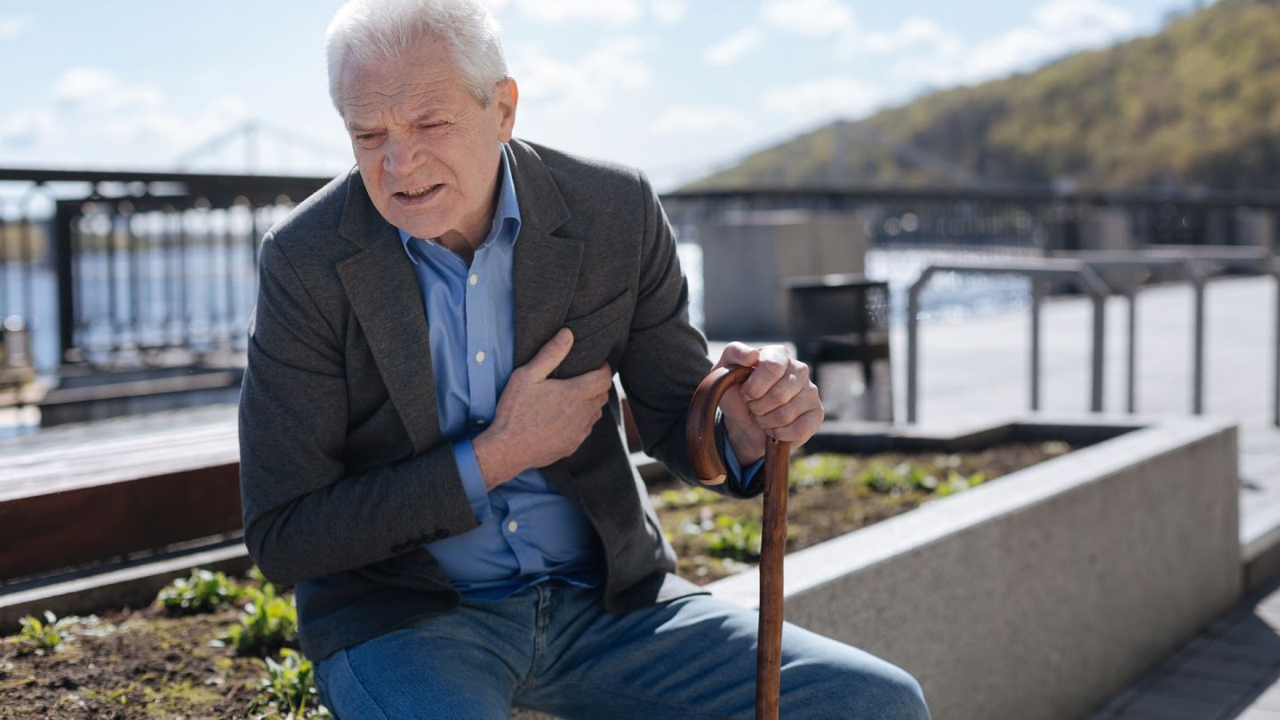 Herzschwaeche (Quelle: Getty Images / Zinkevych) Herzschwäche: Ein älterer Mann sitzt auf einem Stein und hält sich mit einer Hand sein Herz. Sein Gesicht ist angespannt und schmerzverzerrt.