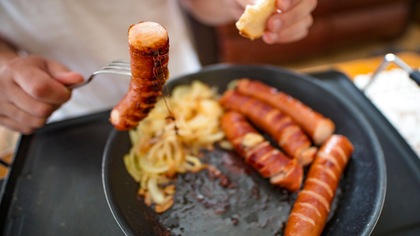 Fettiges Essen (Quelle: Getty Images/mikroman6) Hypercholesterinämie: Ein Mann hält mit einer Gabel eine fettige Wurst. Darunter eine Pfanne.