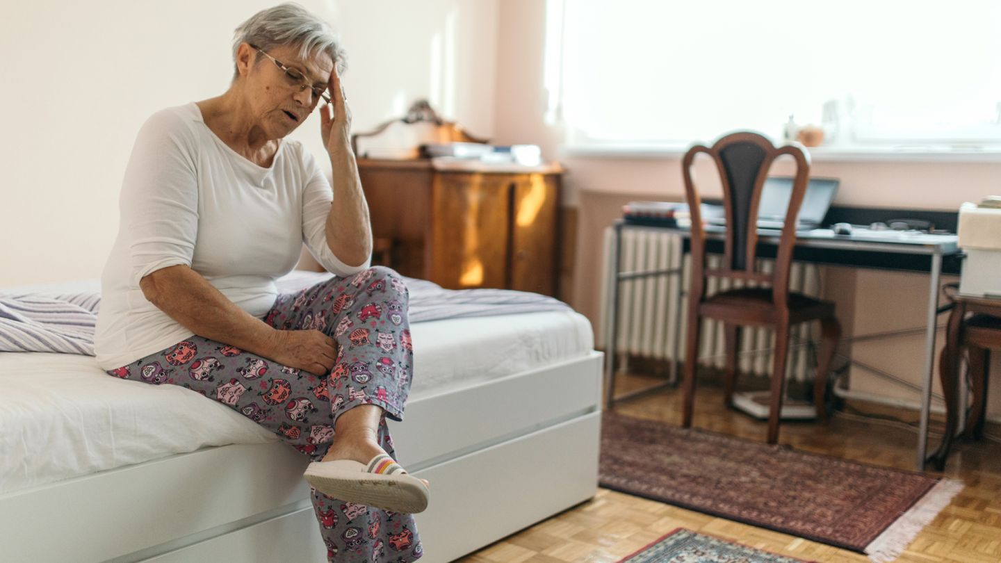 Schwindelgefühl (Symbolbild) (Quelle: Getty Images/supersizer) Lagerungsschwindel: Eine ältere Frau sitzt auf einem Bett, beugt ihren Oberkörper leicht nach vorne und fasst sich mit der linken Hand an die Stirn. Die Frau hat ihren Mund leicht geöffnet. Sie wirkt erschöpft.