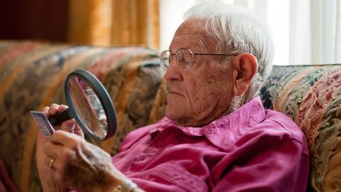 Mann mit Lupe (Quelle: Getty Images/DaveMcDPhoto) Ein älterer Mann sitzt auf einem Sessel. Er hält eine große Lupe in der Hand und blickt durch sie auf eine Plastikkarte.