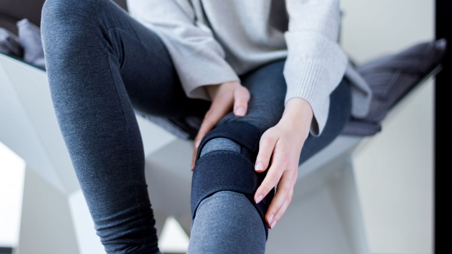 Bein mit Stützbandage (Quelle: Getty Images/monstArrr_) Meniskusriss: Eine Frau sitzt auf einem Stuhl, ihr linkes Bein ist ausgestreckt. Um ihr linkes Knie ist eine Stützbandage gebunden.