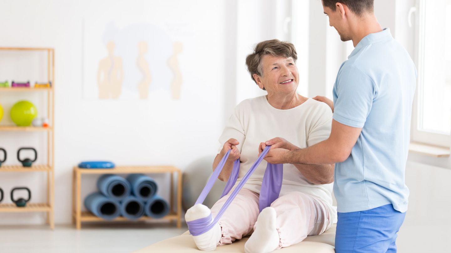 Gymnastik (Quelle: Getty Images/KatarzynaBialasiewicz) Osteoporose: Eine ältere Fau sitzt mit augestreckten Beinen auf einer Isomatte. Um einen Fuß hat die Seniorin ein Fitnessband gespannt, an dem sie mit beiden Händen zieht. Ein Trainer steht neben der Frau und stützt sie am Rücken und an einer Hand.