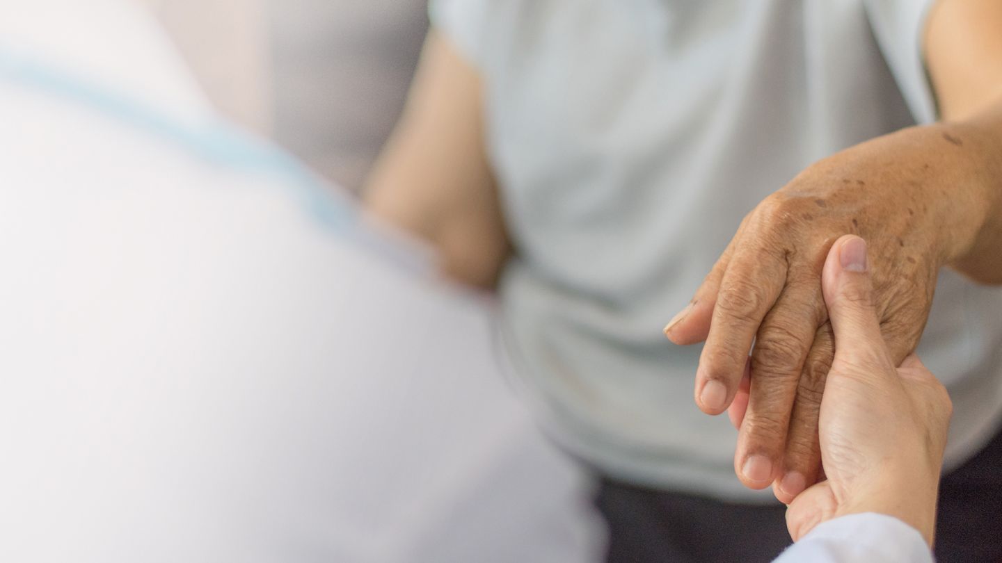 Betreuung bei Parkinson (Quelle: Getty Images/Pornpak Khunatorn) Parkinson: Eine Frau in einem weißen Kittel, offenbar eine Pflegerin, nimmt eine ältere Frau an beide Hände.