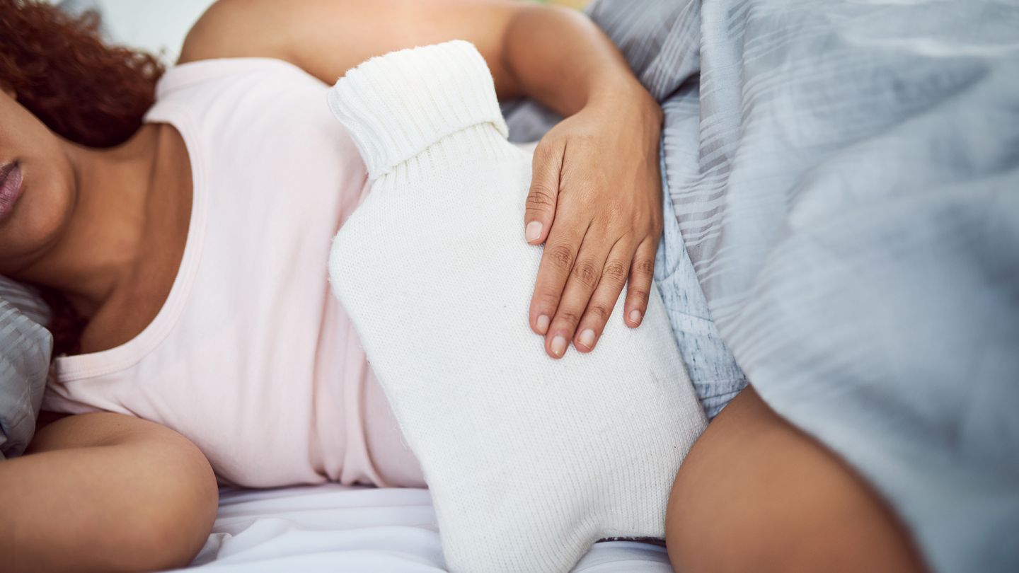 Prämenstruelles Syndrom (PMS) (Quelle: Getty Images/LaylaBird) Prämenstruelles Syndrom (PMS): Eine Frau liegt im Bett und hält sich eine Wärmeflasche auf den Bauch.
