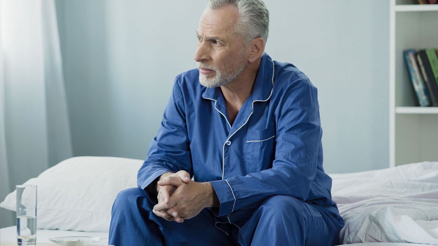 Mann in ärztlicher Behandlung (Symbolbild) (Quelle: Getty Images/Motortion) Gutartige Prostatavergrößerung (Prostatahyperplasie): Ein Mann mittleren Alters sitzt am Rand eines Krankenbettes. Er hat seine Hände ineinander gefaltet und schaut nachdenklich in Richtung Fenster.
