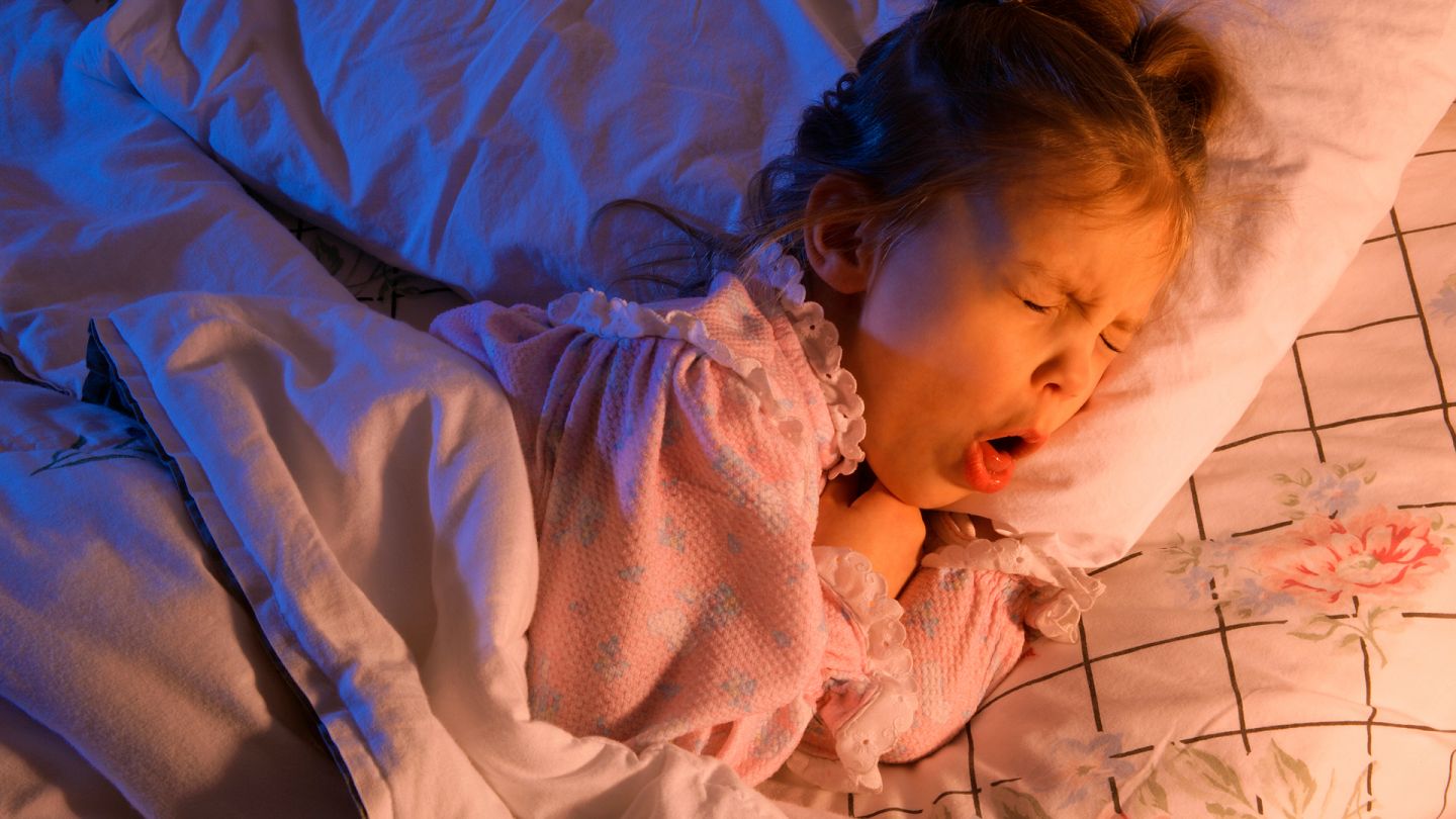 Mädchen mit Husten (Quelle: Getty Images/Design Pics/Don Hammond) Pseudokrupp: Ein kleines Mädchen liegt in einem Bett. Sie hat sich leicht auf die Seite gedreht, den Mund weit geöffnet und hustet stark.