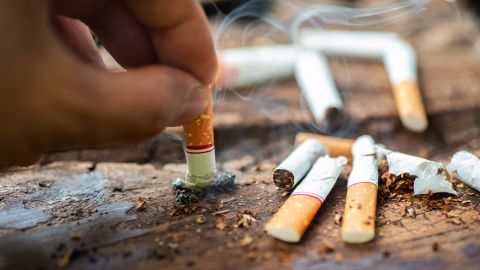 Zigarettenstummel (Quelle: Getty Images/krisanapong detraphiphat) Rauchen: Eine Hand hält eine qualmende Zigarette und drückt sie auf einem hölzernen Untergrund aus. Ringsherum liegen einige Zigarettenstummel.