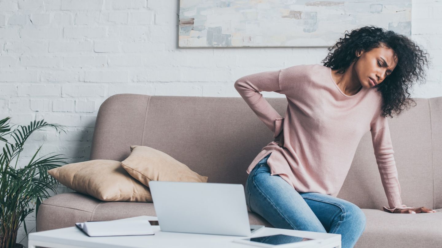 Rückenschmerzen (Quelle: Getty Images/LightFieldStudios) Rückenschmerzen: Eine junge Frau sitzt auf einer Couch und hat ihren Oberkörper leicht zur Seite gebeugt. Mit einem Arm stützt sich die Frau ab, mit der anderen Hand fasst sie sich an den Rücken.