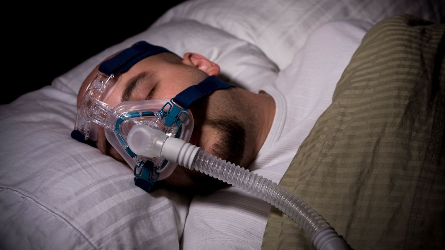 Schlafapnoe (Quelle: Getty Images/rdegrie) Schlafapnoe: Ein Mann liegt in einem Bett und trägt eine Beatmungsmaske. Er scheint zu schlafen.