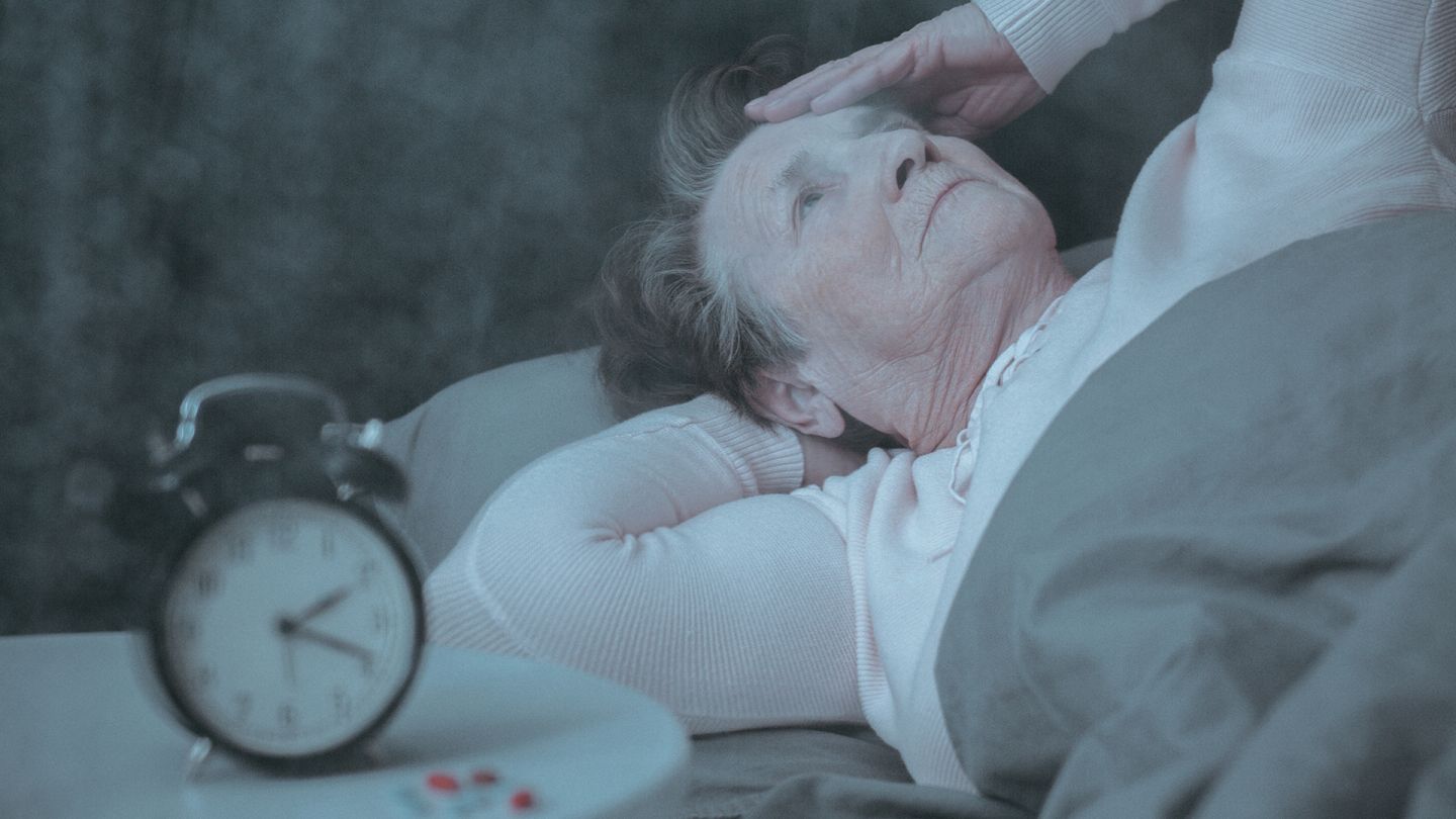 Schlaflos (Symbolbild) (Quelle: Getty Images/KatarzynaBialasiewicz) Schlafstörungen: Eine ältere Frau liegt im Bett, fasst sich an den Kopf und schaut an die Decke – neben ihr auf dem Nachttisch steht ein Wecker.