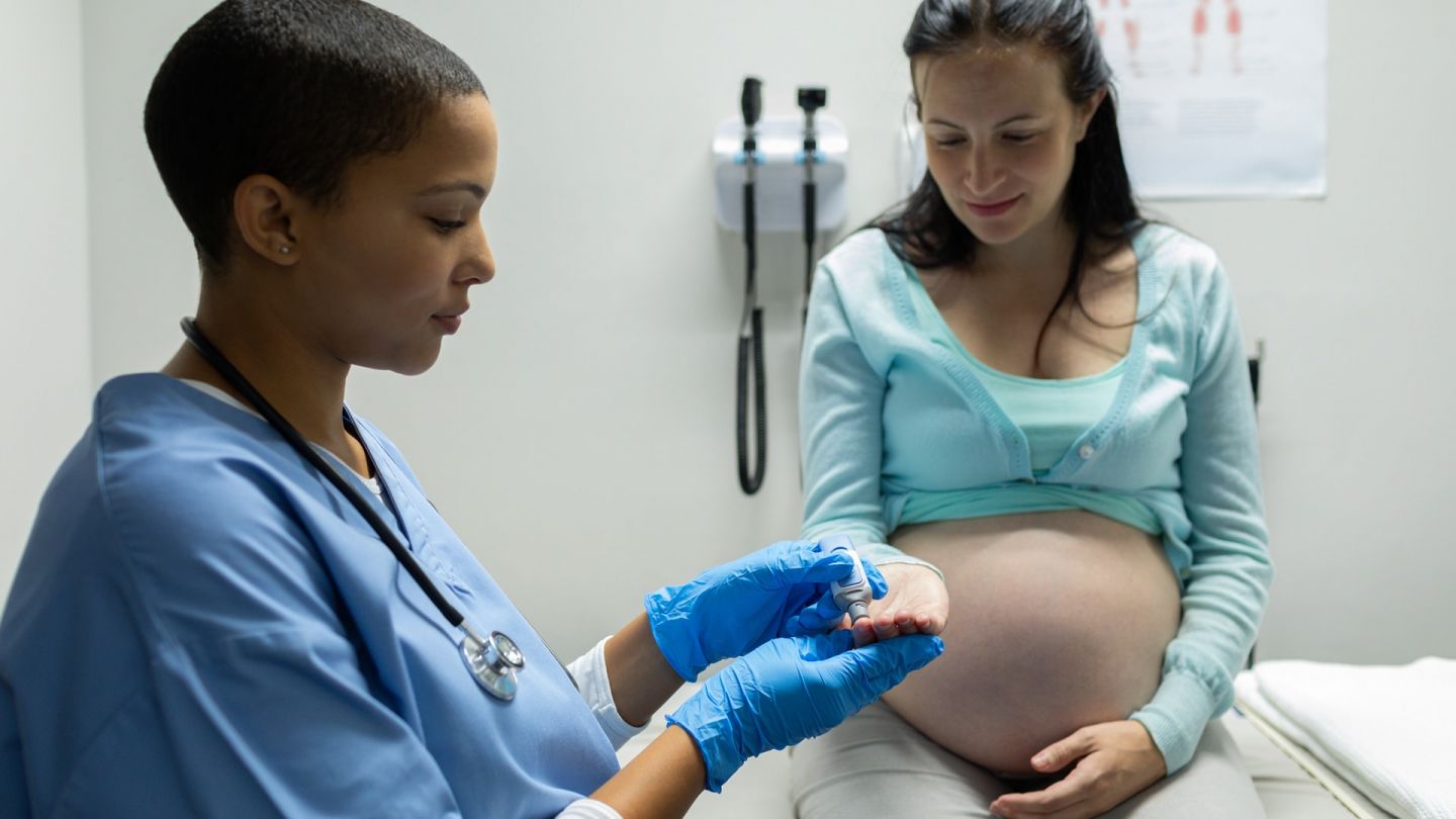 Schwange Frau in der Arztpraxis (Quelle: Getty Images/Wavebreakmedia) Schwangerschaftsdiabetes: Eine schwangere Frau sitzt auf der Behandlungsliege. Neben ihr steht eine Frau, die einen blauen Kittel und Medizinhandschuhe trägt. Sie hält ein Messgerät an die ausgestreckte Hand der Schwangeren.