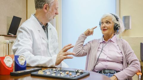 Frau trägt Kopfhörer beim Ohrenarzt (Quelle: GettyImages/Halfdark) Eine ältere Frau sitzt neben einem Hörgeräte-Akustiker und trägt einen Kopfhörer. Sie schaut den Mann an und gestikuliert mit einer Hand. Vor ihnen auf einem Tisch liegen nebeneinander ausgebreitet unterschiedliche Hörgeräte, Hörhilfen und Otoplastiken.