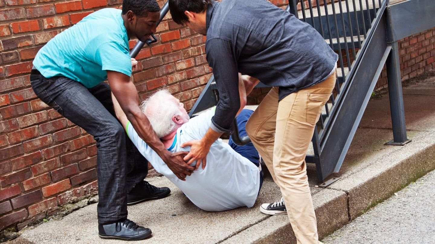 Gestürzter Senior (Quelle: Getty Images/brittak) Stürze bei älteren Menschen: Zwei Männer stehen vor einer Treppe und stützen mit ihren Händen einen älteren Mann, der mit seinem Gesäß auf einem Gehweg sitzt und seinen Oberkörper nach vorne gebeugt hat. Die Beine des Senioren berühren die Treppenstufen.