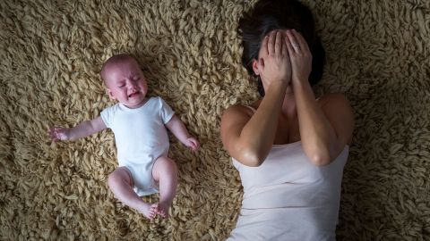 Depressive Mutter mit Kind (Quelle: Getty Images/SolStock) Eine Frau liegt auf einem Teppich und hält sich beide Hände vors Gesicht. Sie wirkt erschöpft. Neben ihr liegt ein Baby. Es scheint zu strampeln und zu weinen.