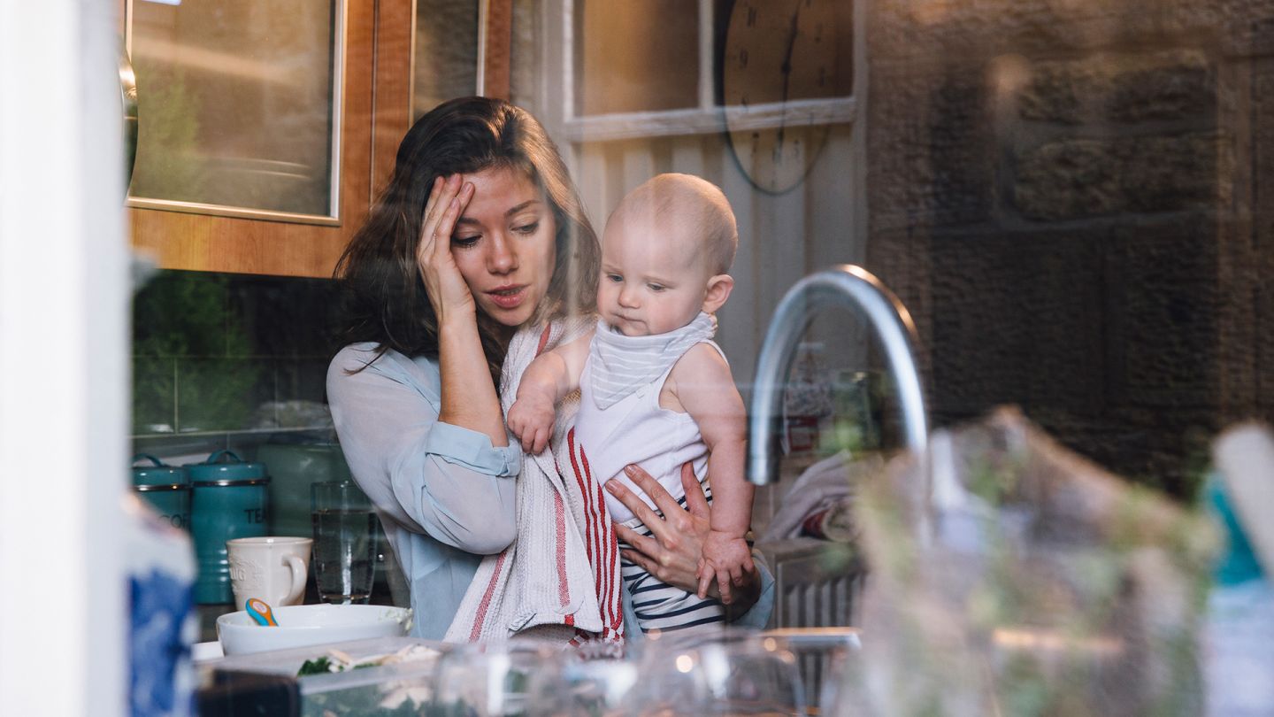 Mutter mit Baby (Quelle: Getty Images/SolStock) Wochenbettdepression: Eine gestresst wirkende Frau steht in der Küche, hält ein Baby im Arm und hält sich den Kopf.